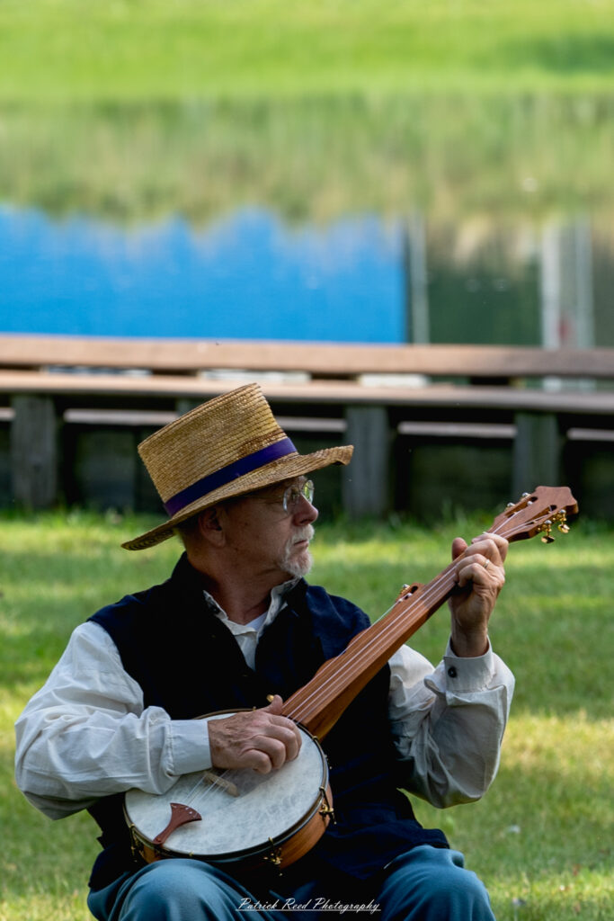 A seated man playing a banjo, his fingers skillfully strumming the strings. He is dressed in casual attire, with a relaxed expression that reflects his enjoyment of the music. The setting is warm and inviting, highlighting the joyful atmosphere of a musical performance.